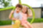 Two smiling children sit on a green playground ring, laughing outdoors in bright daylight.