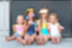 Four smiling girls in swimsuits sit on pavement blowing bubbles, with a black metal wall behind them.