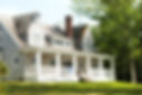 “Classic two-story residential home with gray shingles, surrounded by lush greenery and featuring an expansive white porch.