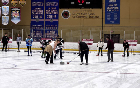 A man and woman sweep the ice ahead of a curling stone while they're watched and cheered on by teammates and other participants.