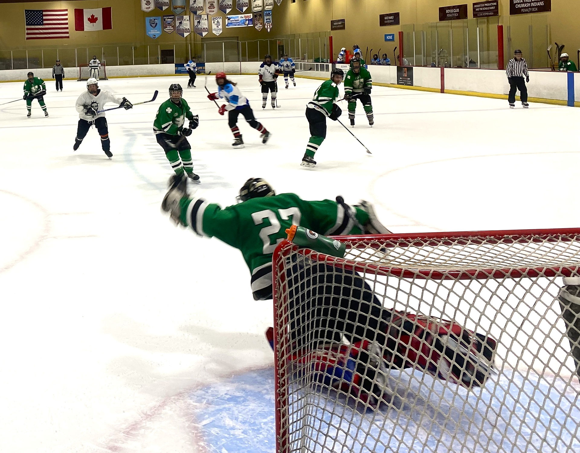 A goalie lunches to block the puck as adult hockey players look on to see the outcome of the shot