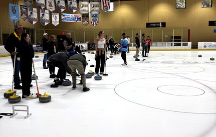 A group of participants prepare to try curling, standing on the ice with their brooms, stones, and other material as they listen to an instructor out of frame.