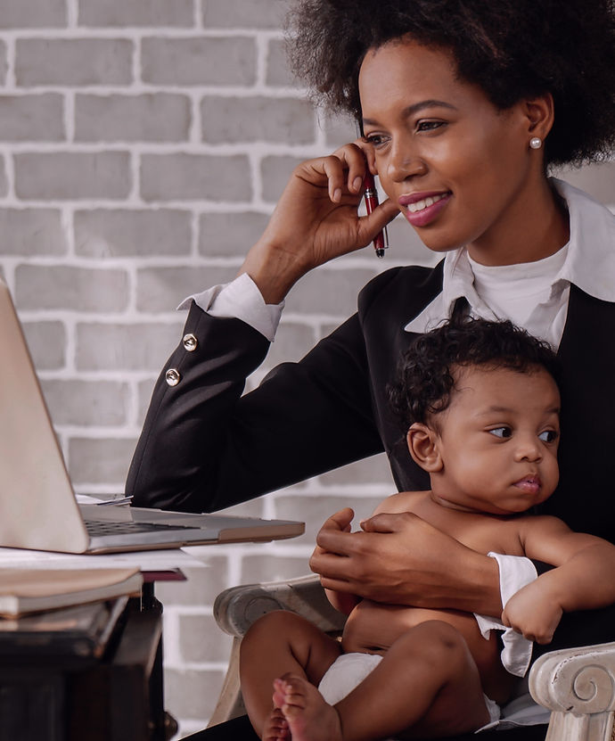 Mulher negra, de cabelos crespos, roupa social, olhando para a tela de um notebook com seu bebê no colo.