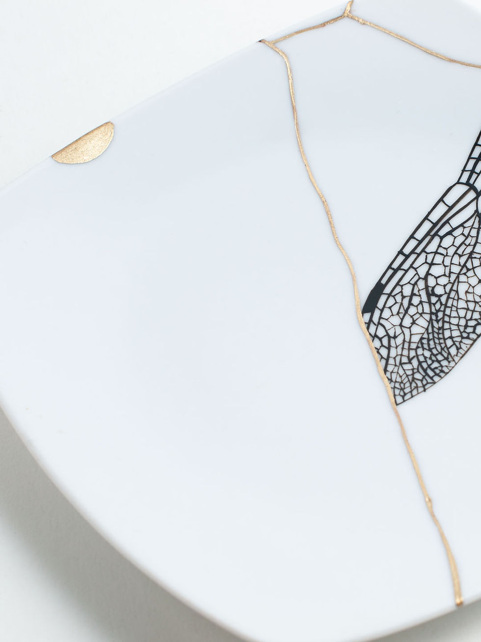 Thumbnail: Kintsugi-KerDetail von amikteller in Weiß. Schwarzes, handgemaltes Flügeldesign auf einer Scherbe, Brüche in Gold ausgeführt.