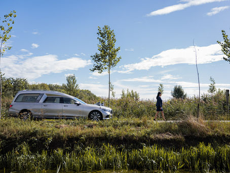 Uitvaartbegeleider van Kamsteeg Uitvaartzorg loopt voor een grijze rouwauto in natuurlijke omgeving