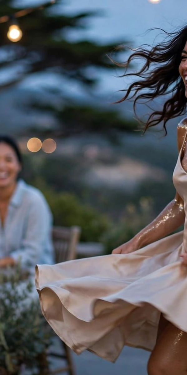 A woman in a champagne satin dress twirls on a terrace strung with lights, friends gathered at a long table behind her.