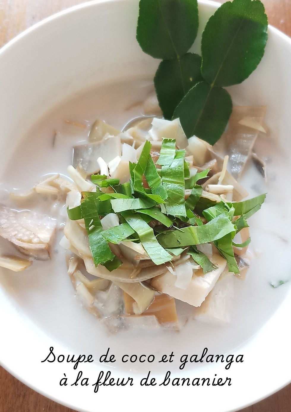 Bol de soupe de coco et galanga à la fleur de bananier, garni de feuilles de kaffir fraîches. Une soupe crémeuse aux arômes thaïlandais, mettant en valeur la texture délicate de la fleur de bananier.