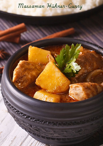 Eine Schale Massaman Hühner-Curry mit saftigem Hähnchen, goldgelben Kartoffeln und einer würzigen Kokosmilch-Soße, garniert mit frischen Kräutern. Im Hintergrund sind Zimtstangen und Sternanis zu sehen, die dem Gericht sein unverwechselbares Aroma verleihen.