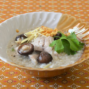 Bol de khao tom pla, soupe de riz thaïlandaise au poisson, garni de champignons shiitake, gingembre en julienne, ail frit et feuille de céleri, servi dans un bol sur une nappe thaïlandaise colorée.