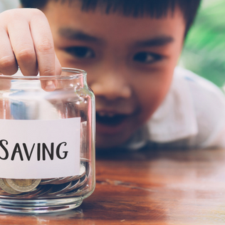 A child is smiling while putting a coin into a glass jar labeled "SAVING."