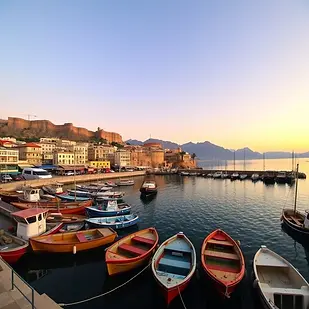 A wide-angle photo of Kyrenia Harbour at sunrise, showing colourful fishing boats, the curved harbour wall, and Kyrenia Castle glowing in the early morning light, with the Besparmak Mountains rising softly in the background.