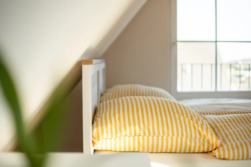 2 Kopfkissen mit gelb-weißen Bezug, weißes Bett, im Hintergrund ein Fenster