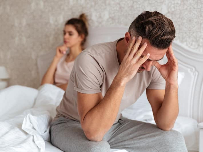 Upset man sitting on bed, woman in background