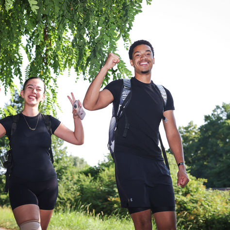 Zwei lächelnde Wanderer beim Megamarsch in schwarzer Sportkleidung zeigen Peace-Zeichen im grünen Park unter Baumästen.