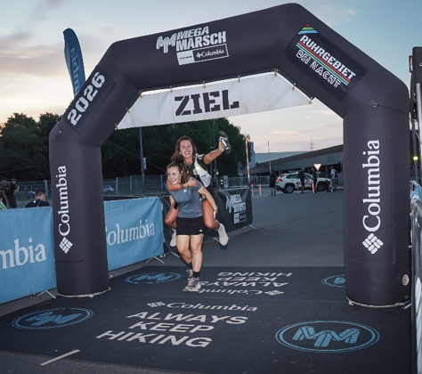Zwei Frauen laufen lachend durchs Ziel unter einem Columbia-Arc mit ZIEL., bei dem Megamarsch in Abendstimmung.
