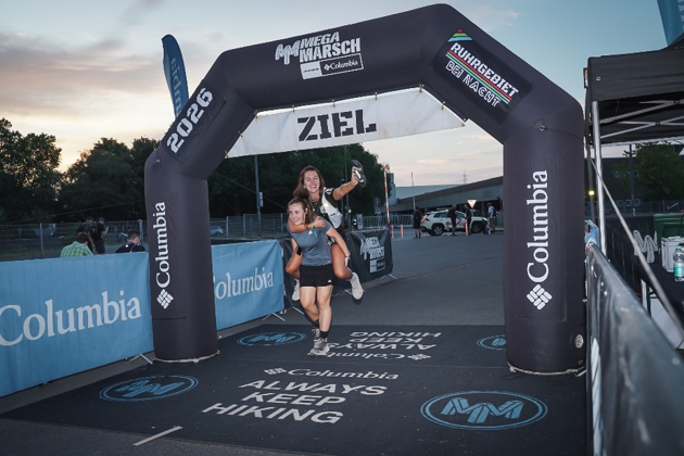 Zwei Frauen laufen lachend durchs Ziel unter einem Columbia-Arc mit ZIEL., bei dem Megamarsch in Abendstimmung.