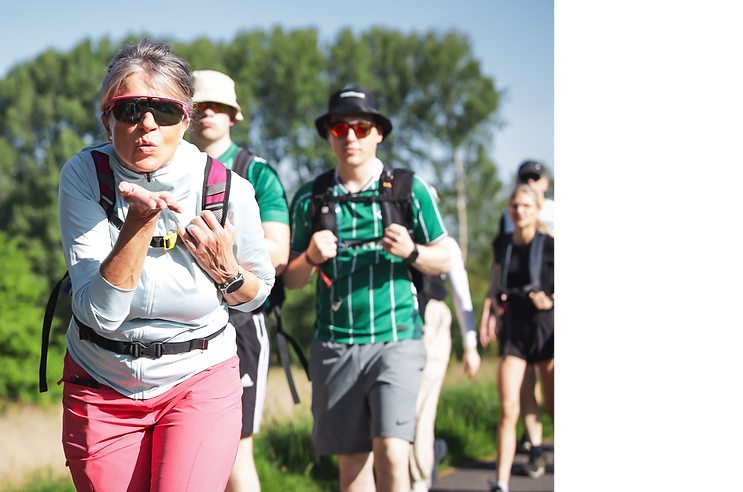 Teilnehmende beim Megamarsch auf einer Wanderstrecke durch die Natur – eine Frau schickt motiviert einen Kuss in die Kamera, während die Gruppe gemeinsam weitergeht.