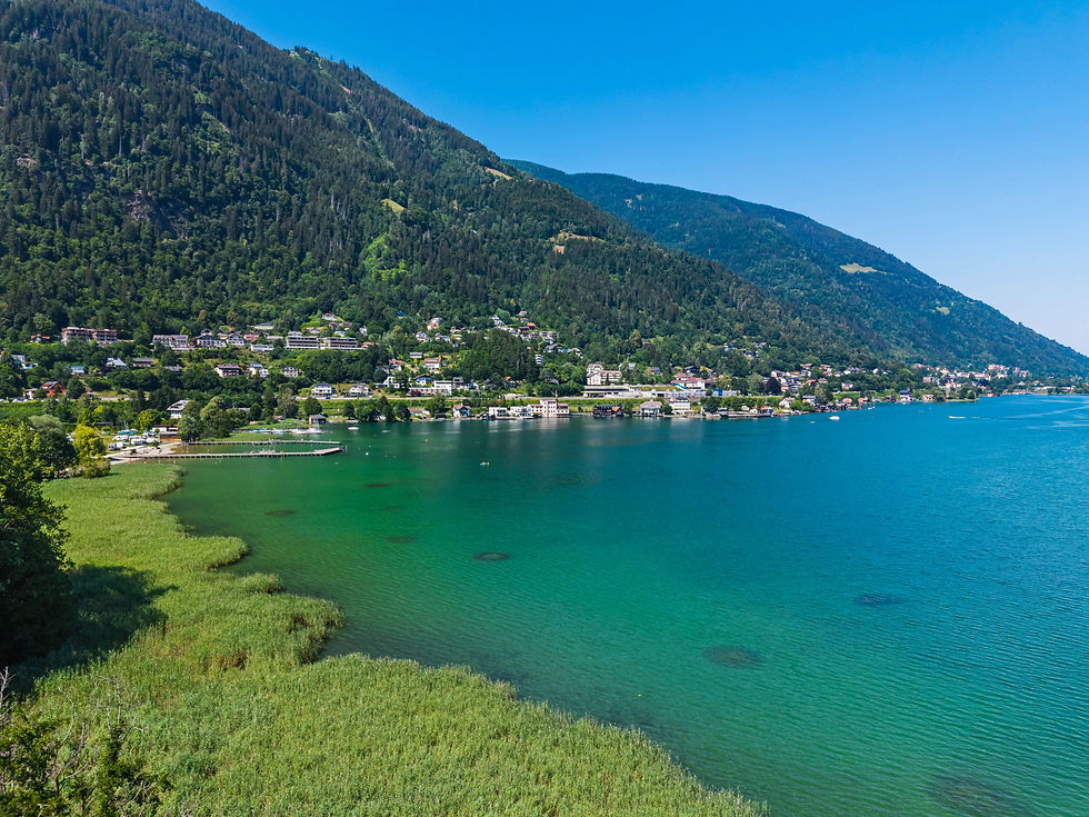 Ossiacher See mit Uferdorf am Fuß bewaldeter Berge unter blauem Himmel.