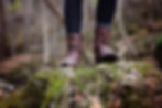 Person mit braunen, geschnürten Stiefeletten steht auf einem moosbewachsenen Felsen in einem herbstlichen Wald, umgeben von Bäumen und Laub.
