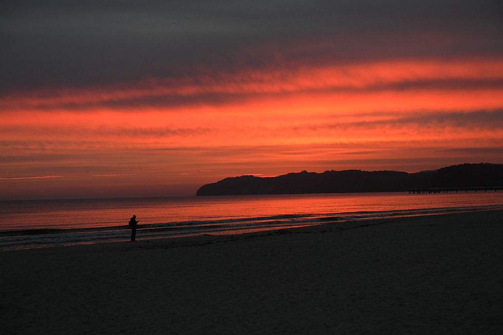 Silhouette einer Person am Strand bei rotem Sonnenuntergang über dem Meer, dunkle Küste im Hintergrund.