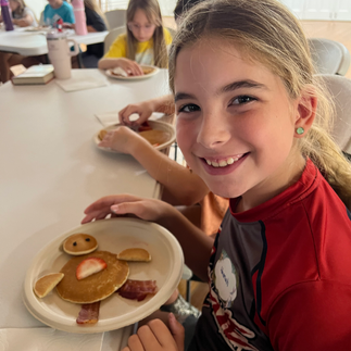 A girl smiling with a plate in front of her holding pancake pieces arranged to look like a penguin