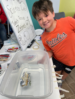 A boy standing next to a bin of water where he has placed a homemade boat made of tin foil, sticks, and straws.