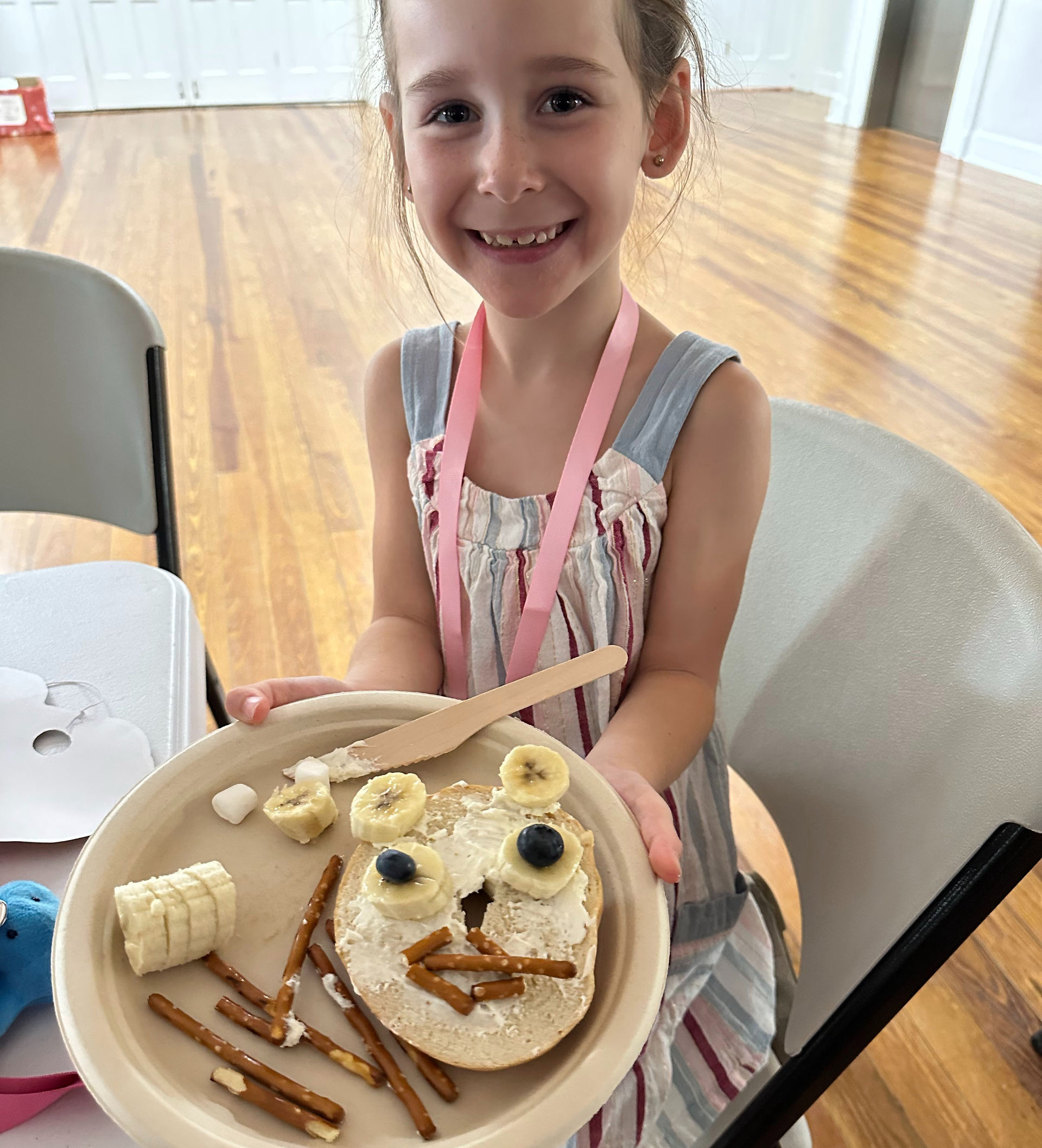 A girl with a bagel on a plate, with bannas and blueberries and pretzels to make an animal face
