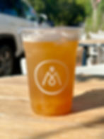 A clear plastic cup of iced peach tea with visible fruit pulp at the bottom, served with a lid and featuring the minimalist white logo of a local café. The drink is placed on a wooden outdoor table with a sunny backdrop, including a parked car and shaded seating area.