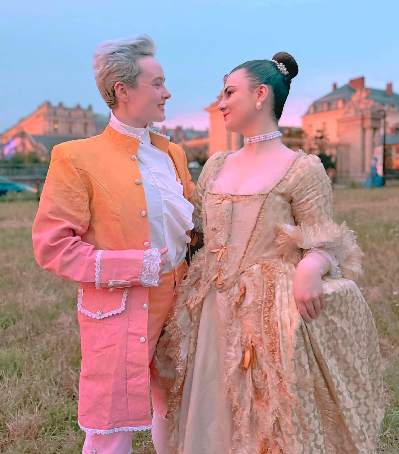 Two attendees in coordinating peach and pink Rococo costumes pose together at sunset before the Versailles Grand Masked Ball masquerade.