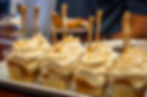 Individual servings of peach dessert in clear plastic cups, each layered with vanilla cake, peach compote, coconut cream diplomat, and topped with peach buttercream and toasted coconut flakes. Each cup is garnished with a gold spoon and arranged on a serving tray.