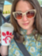 A woman wearing cream-colored sunglasses and a floral blue-green blouse smiles while holding a Chick-fil-A peach milkshake. She is seated in a car with a seatbelt on, enjoying the sunny day through the car window.