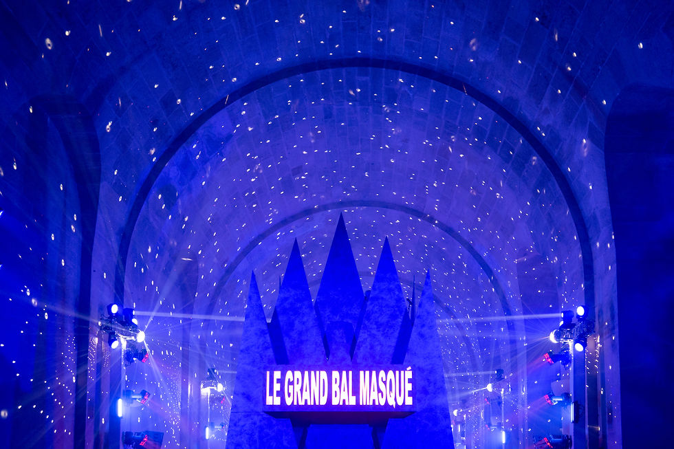 Illuminated entrance inside the Orangerie at the Versailles Grand Masked Ball, featuring dramatic blue lighting, a crown-shaped stage backdrop, and a glowing sign that reads "Le Grand Bal Masqué" beneath a vaulted, starry ceiling.