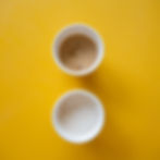 Top-down view of two white paper cups on a bright yellow background. The top cup contains iced coffee with a visible ice cube, while the bottom cup holds a foamy, creamy beverage, possibly a smoothie or latte. The vibrant backdrop contrasts with the neutral tones of the drinks.