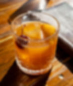 A close-up of a Peach Old Fashioned cocktail served in a cut-glass tumbler. The drink features a large ice cube, an orange peel, and a bourbon-soaked cherry garnish. It rests on a sunlit wooden table with a leather wallet or menu nearby.
