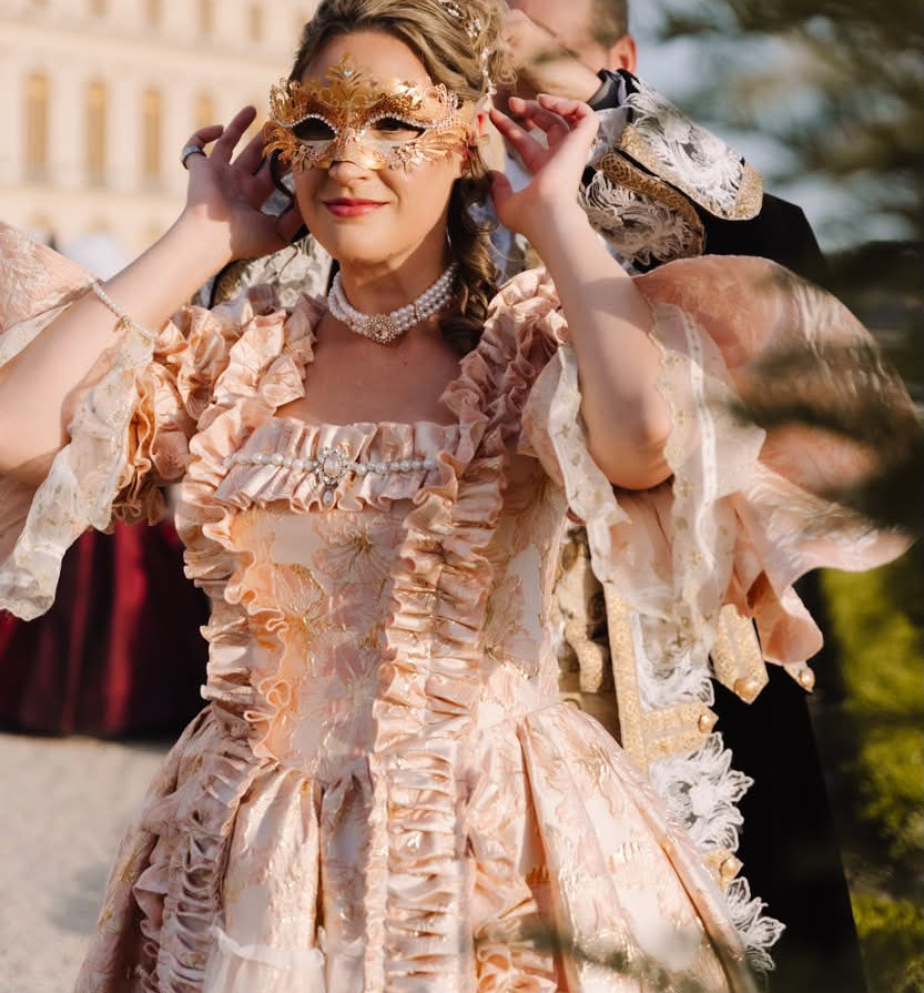A woman wearing an ornate blush-pink Rococo costume adjusts her gold masquerade mask while posing at the Versailles Grand Masked Ball.