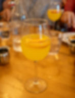 A glass of peach mimosa served in a wine glass, garnished with a thin slice of fresh peach. The drink sits on a wooden table surrounded by water glasses, a glass bottle, and other brunch items, with a blurred background of people dining.