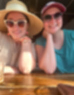 Two women smiling and seated at a wooden table in a sunlit café. The woman on the left wears a wide-brimmed straw hat, oversized sunglasses, peach-shaped earrings, and a cream eyelet blouse. The woman on the right wears a red baseball cap, round sunglasses, and a teal shirt. Both rest their chins on their hands, enjoying a relaxed moment together.