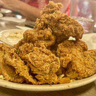 A towering plate of crispy golden garlic fried chicken, served on a bed of cabbage at a restaurant in Hawaii. The crunchy, well-seasoned batter glistens under the light, highlighting the delicious texture. In the slightly blurred background, a diner wearing a red Hawaiian dress and a lei enjoys the meal, adding to the island dining experience.