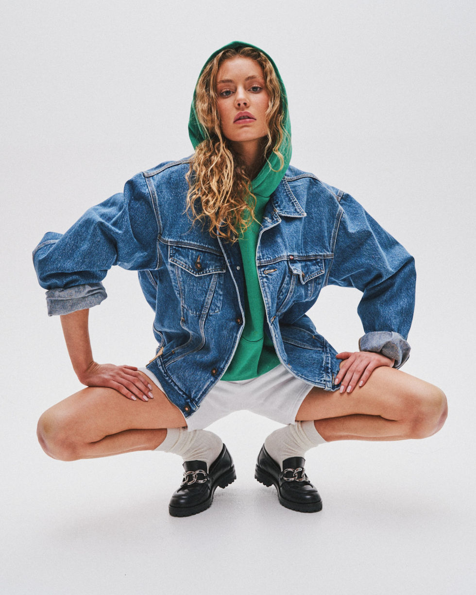 Model in an oversized denim jacket and pale cap crouching against a grey studio backdrop, Melbourne, Australia; styling by Tamarra McNaught, founder of TITO STYLE.
