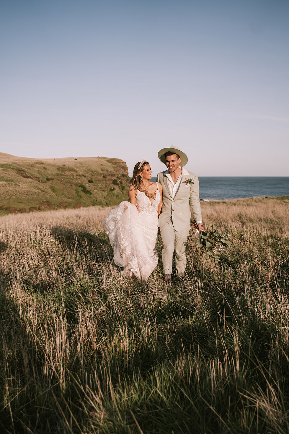 A couple walking through golden grass at dusk. A gown by The Sposa Group with menswear by YSG Tailors, styling by Tamarra McNaught, founder of TITO STYLE, at Cairn Beach, Australia.
