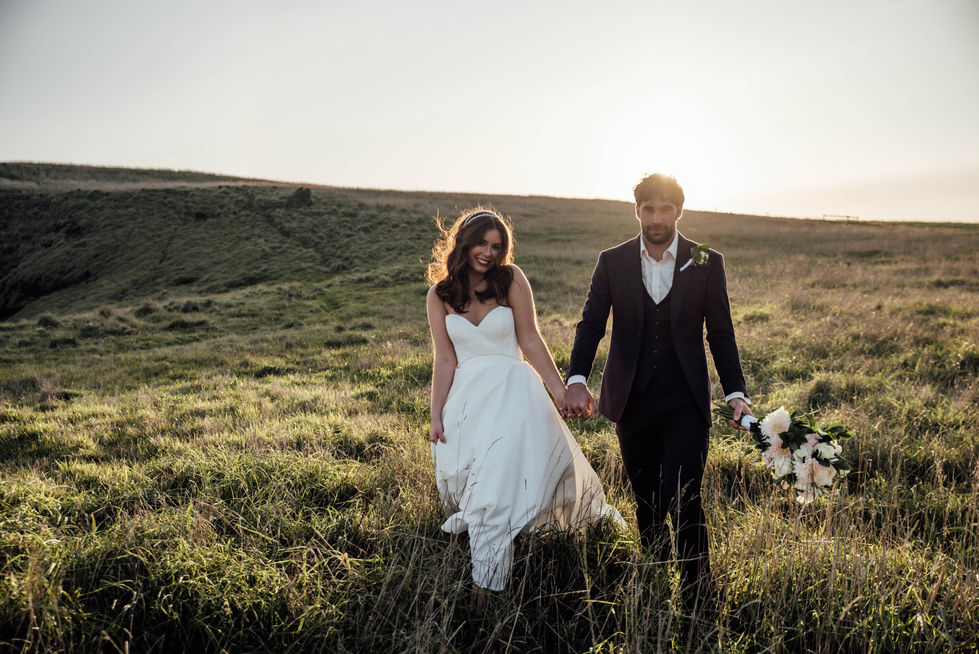 A couple walking hand in hand through a golden field at sunset, the groom in a dark suit. A gown by The Sposa Group with menswear by YSG Tailors, styling by Tamarra McNaught, founder of TITO STYLE, at Cairn Beach, Australia.