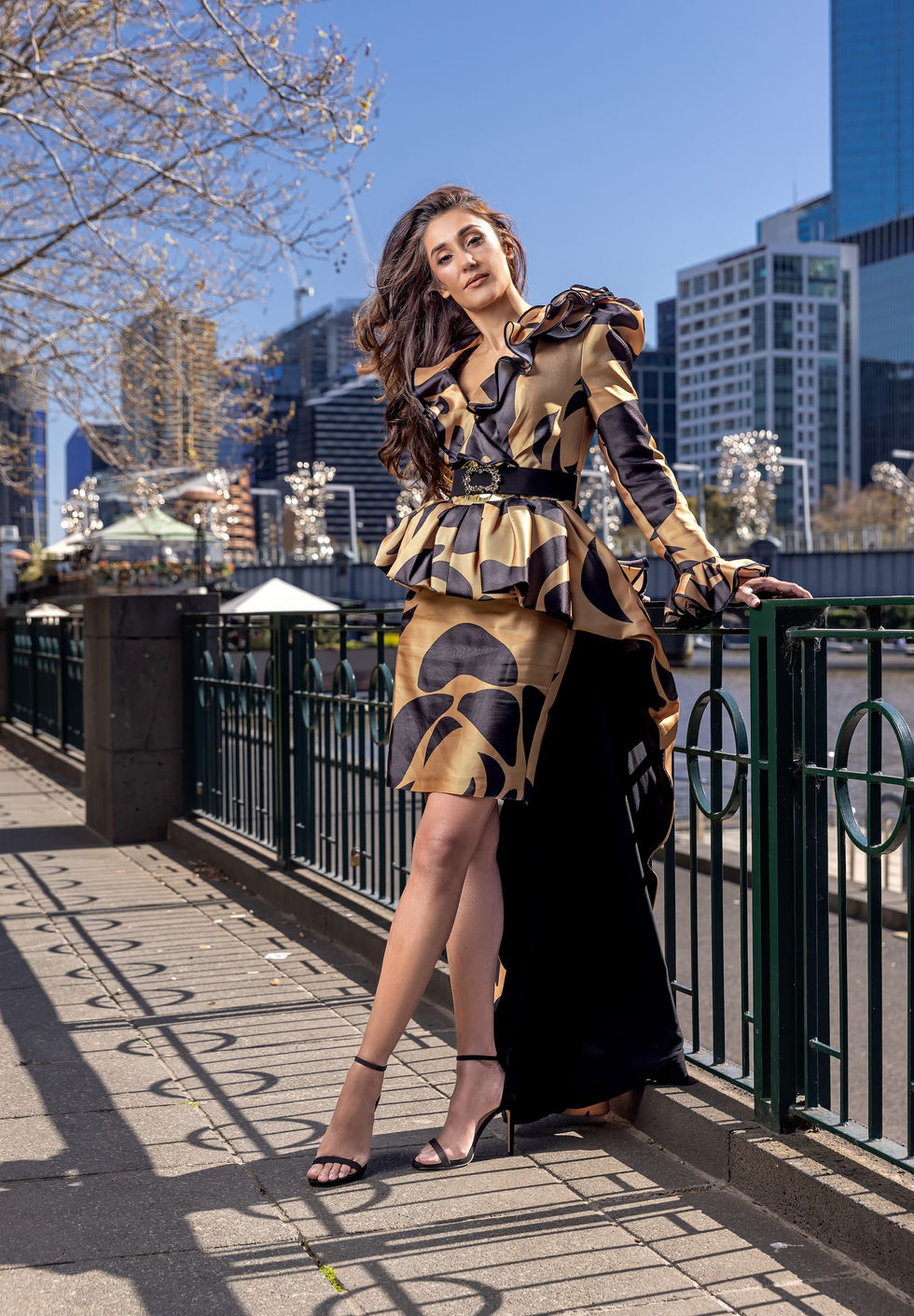 Model in a gold and black Katayoon Mabel ruffled peplum look at the Southbank river railing with the Melbourne skyline behind, Australia; creative direction and styling by Tamarra McNaught, founder of TITO STYLE.