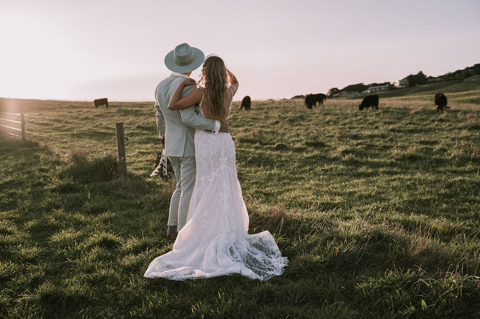 A couple embracing in a field at sunset, the groom in a hat. A gown by The Sposa Group with menswear by YSG Tailors and a hat by Lack of Color, styling by Tamarra McNaught, founder of TITO STYLE, at Cairn Beach, Australia.