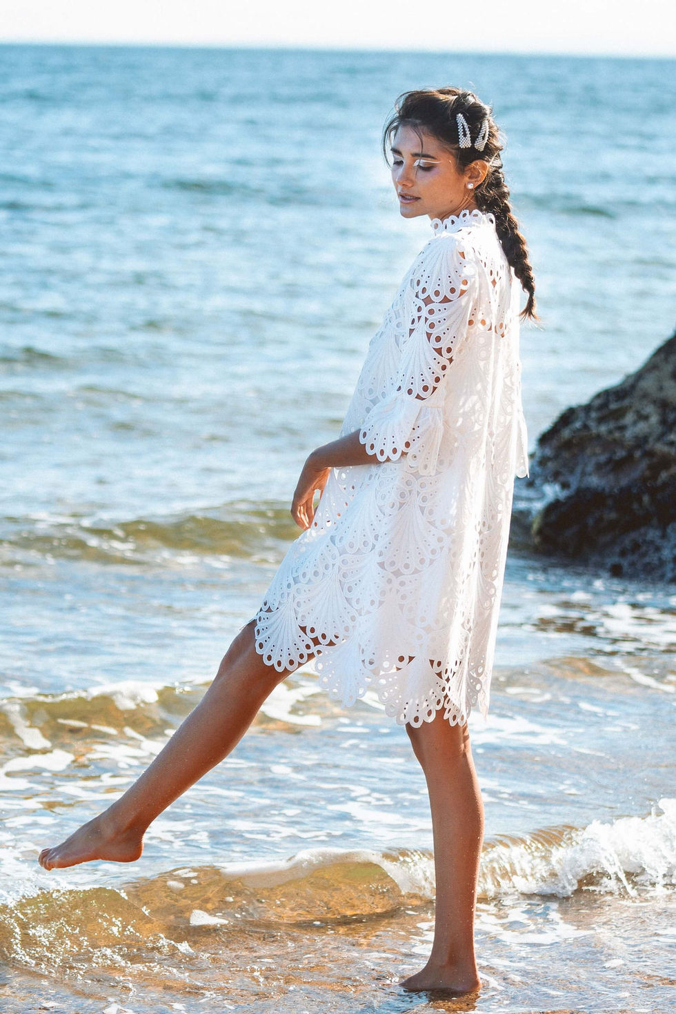 A white broderie anglaise dress at the water's edge at Black Rock, Melbourne, Australia, one foot lifted in the shallows, styling by Tamarra McNaught, founder of TITO STYLE.