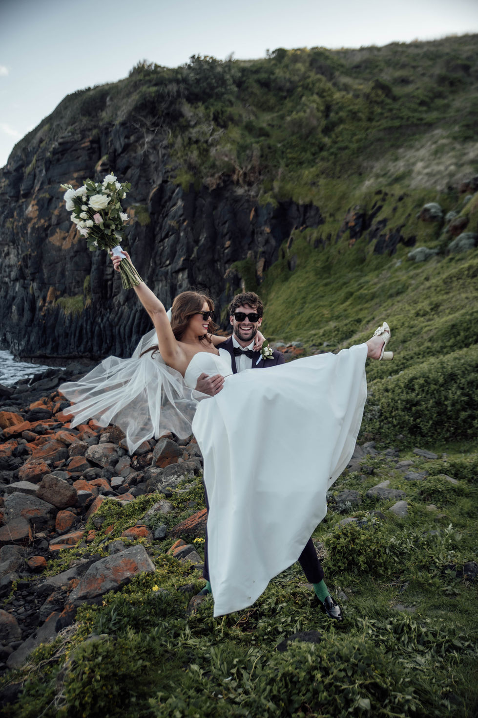 A bride with her veil thrown wide, arms open on the rocks. A gown by The Sposa Group, styling by Tamarra McNaught, founder of TITO STYLE, at Cairn Beach, Australia.