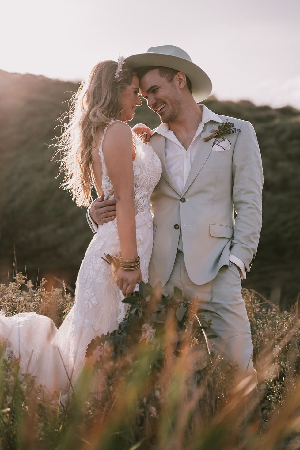A couple close in the long grass, the groom in an olive suit and hat. A gown by The Sposa Group with menswear by YSG Tailors and a hat by Lack of Color, styling by Tamarra McNaught, founder of TITO STYLE, at Cairn Beach, Australia.