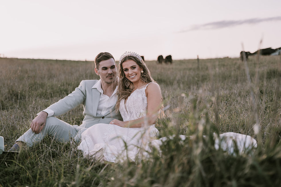 A couple seated together in golden grass, the bride in a floral crown. A gown by The Sposa Group with menswear by YSG Tailors, styling by Tamarra McNaught, founder of TITO STYLE, at Cairn Beach, Australia.