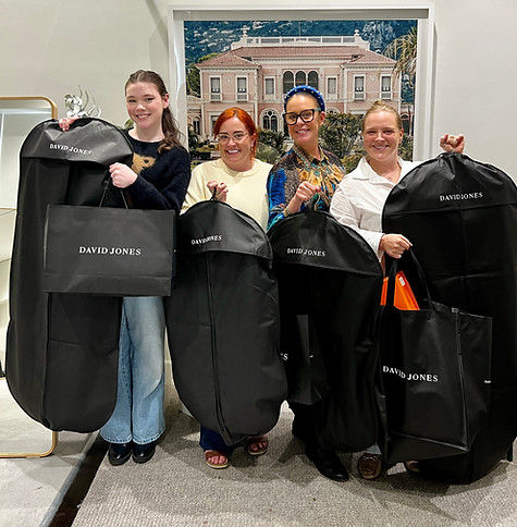 Group of four smiling women proudly holding David Jones and designer garment bags after a successful personal styling session, standing in a luxe fitting suite