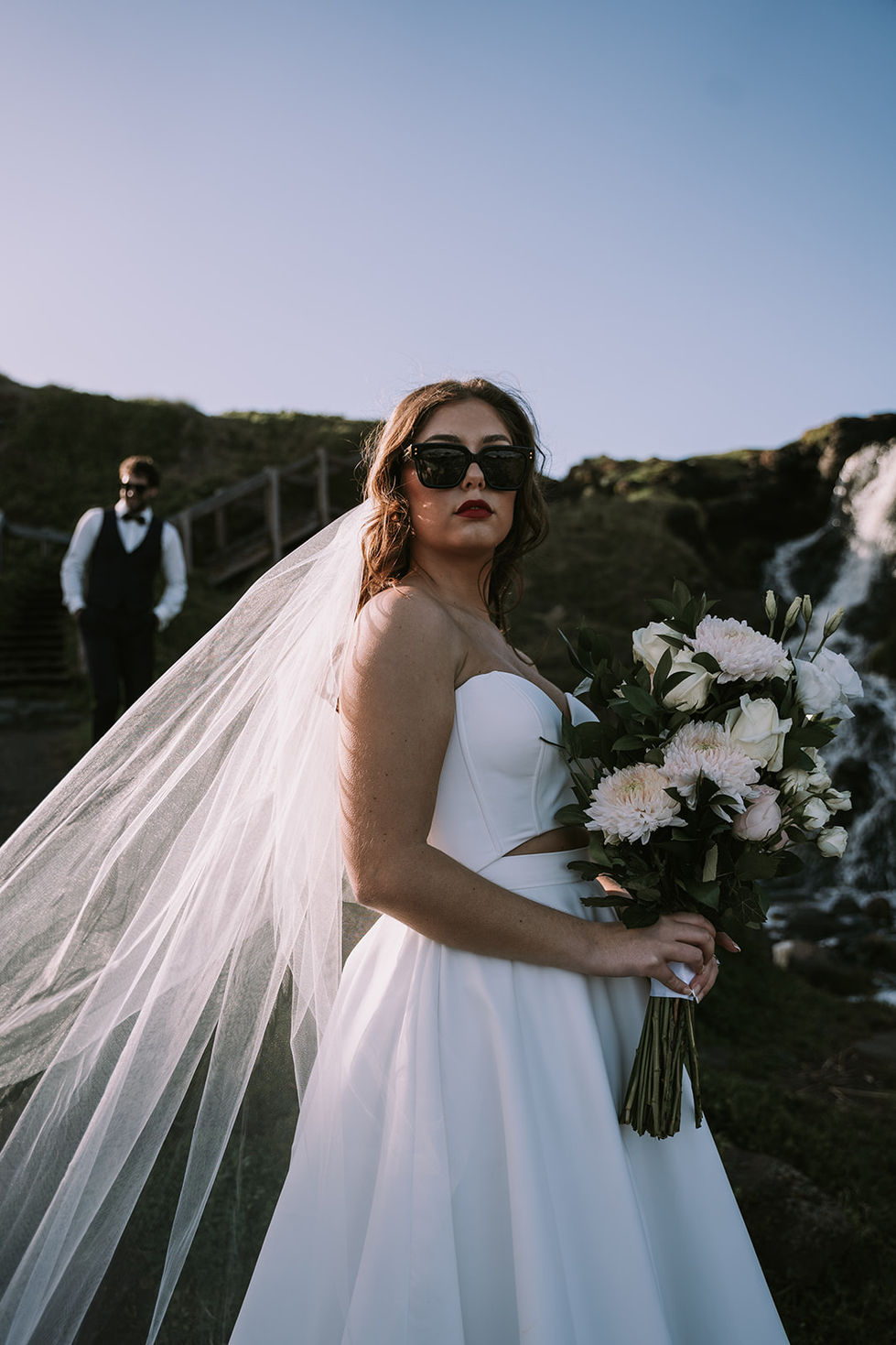 A bride in sunglasses and a veil, holding a bouquet by the waterfall. A gown by The Sposa Group with sunglasses by Gucci, styling by Tamarra McNaught, founder of TITO STYLE, at Cairn Beach, Australia.
