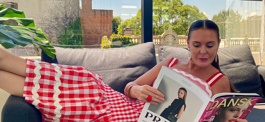 Tamarra McNaught, fashion and celebrity stylist and founder of TITO STYLE, lounges on a sofa in Melbourne, Australia, wearing a red gingham dress while reading a Danish fashion magazine. This candid moment reflects her editorial eye, effortless personal style, and dedication to staying creatively inspired.